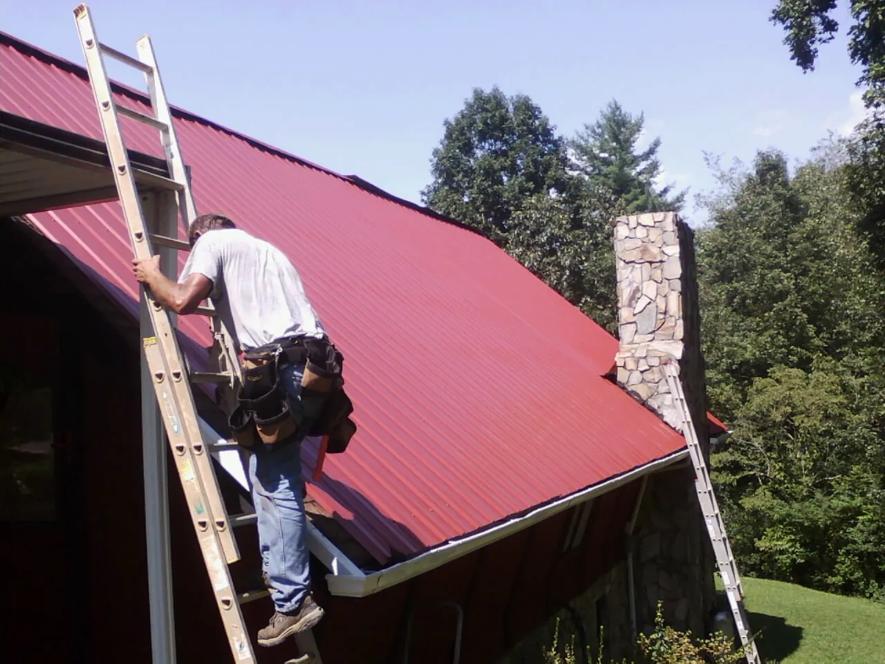 Roofer on a ladder beside a red metal roof and stone chimney — Soffit & Fascia Repair by Roofing Brothers in Perryman